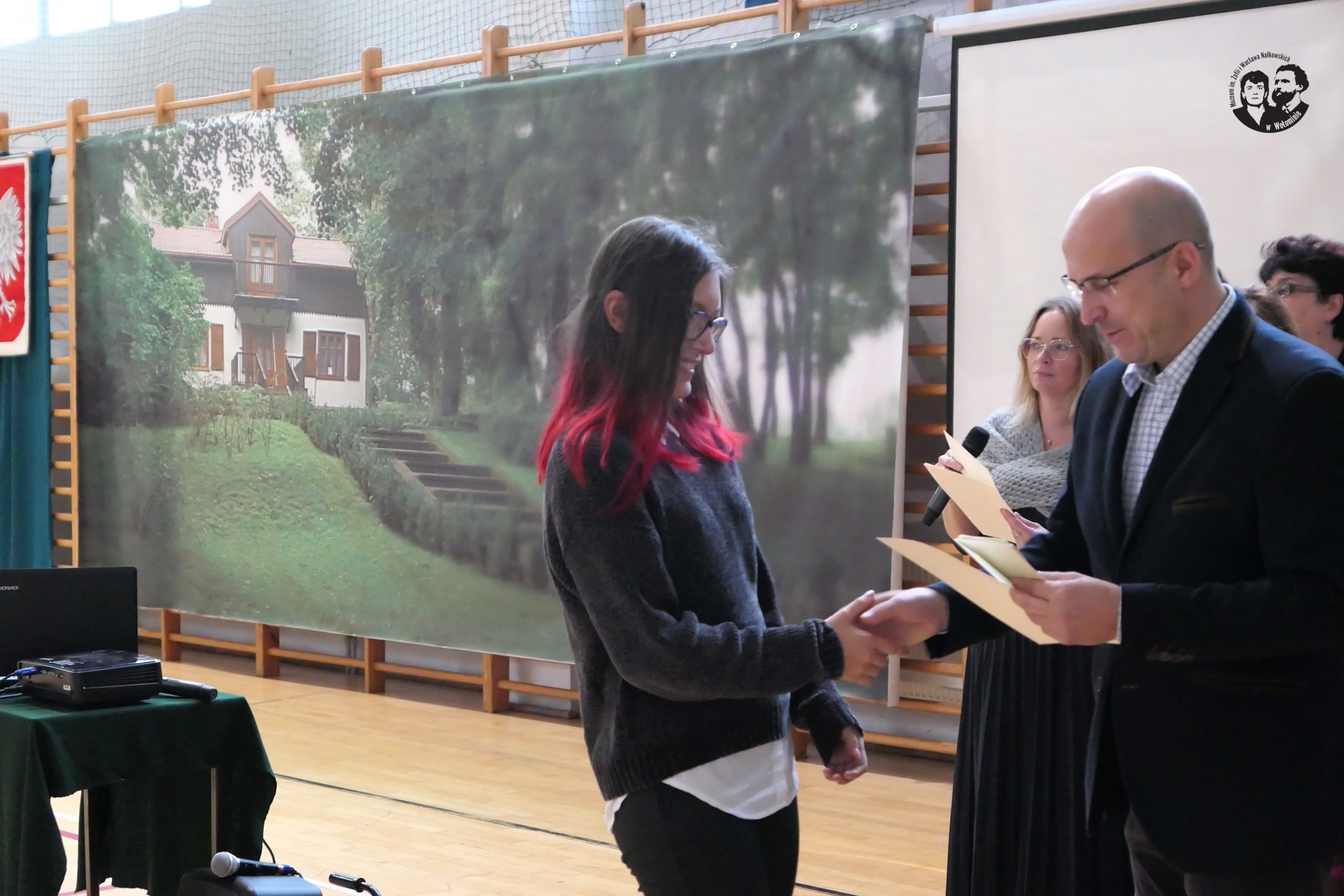 fotografia kolorowa, wnętrze sali gimnastycznej, w tle wizerunek budynku Muzeum, na pierwszym planie z prawej strony wręczający nagrodę Dariusz Szymanowski oraz jedna z laureatek