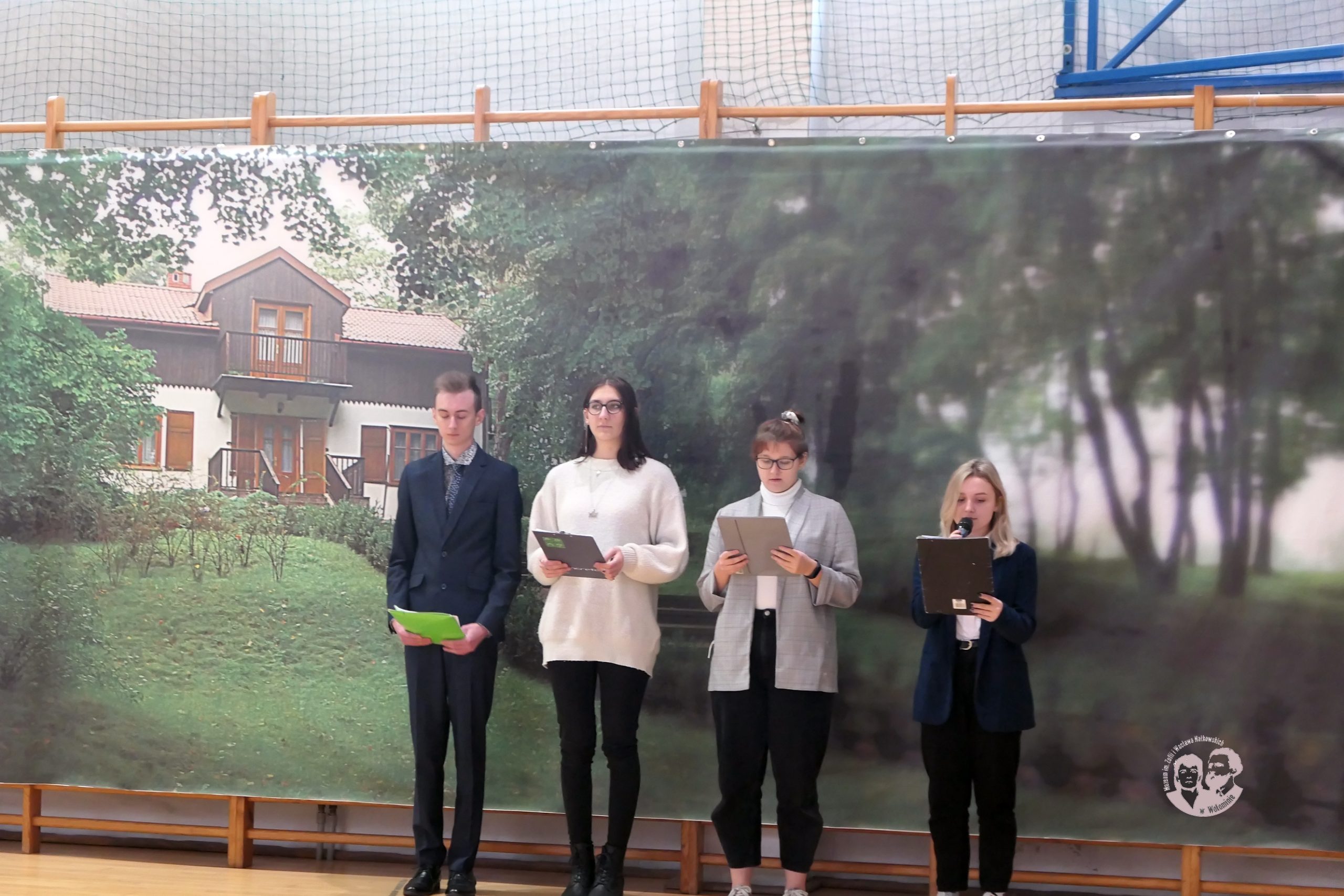 fotografia kolorowa, na tle wizerunku Muzeum czworo przedstawicieli młodzieży (trzy dziewczęta i jeden chłopak), którzy przedstawiają biografię Wacława Nałkowskiego