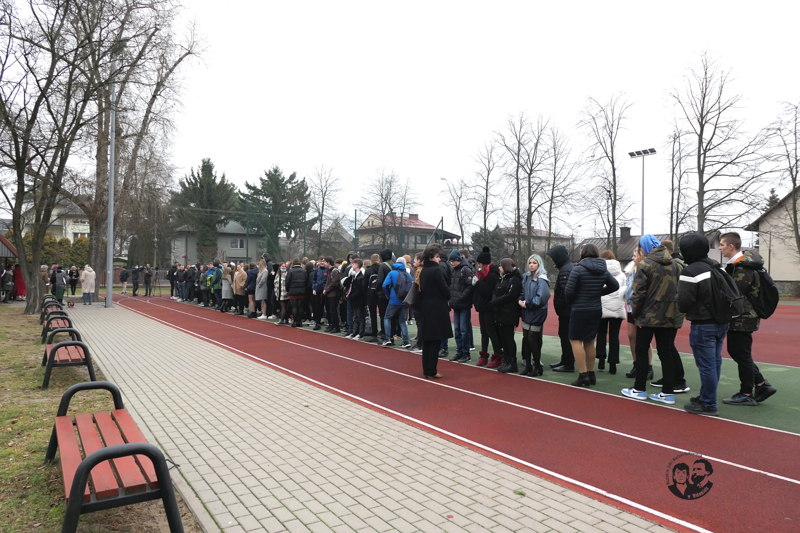 Fotografia kolorowa, na której widać młodzież oczekującą na odsłonięcie muralu, teren sportowy przy szkole.