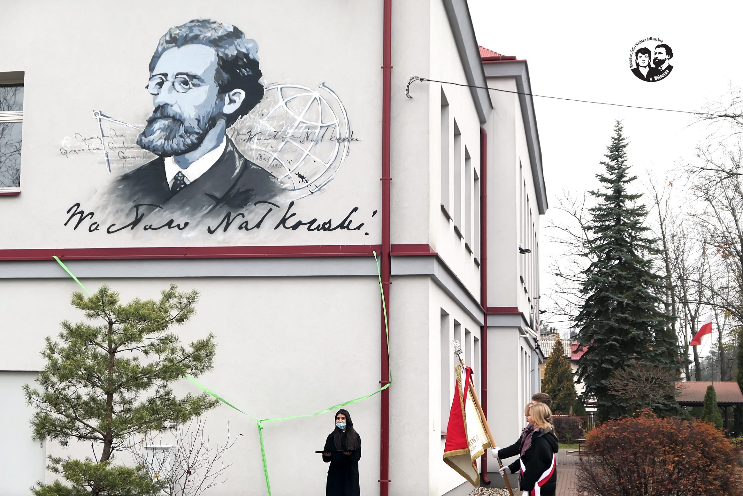 Fotografia kolorowa, przedstawiająca widok ściany budynku z muralem oraz przedstawicielkę młodzieży i poczet sztandarowy szkoły