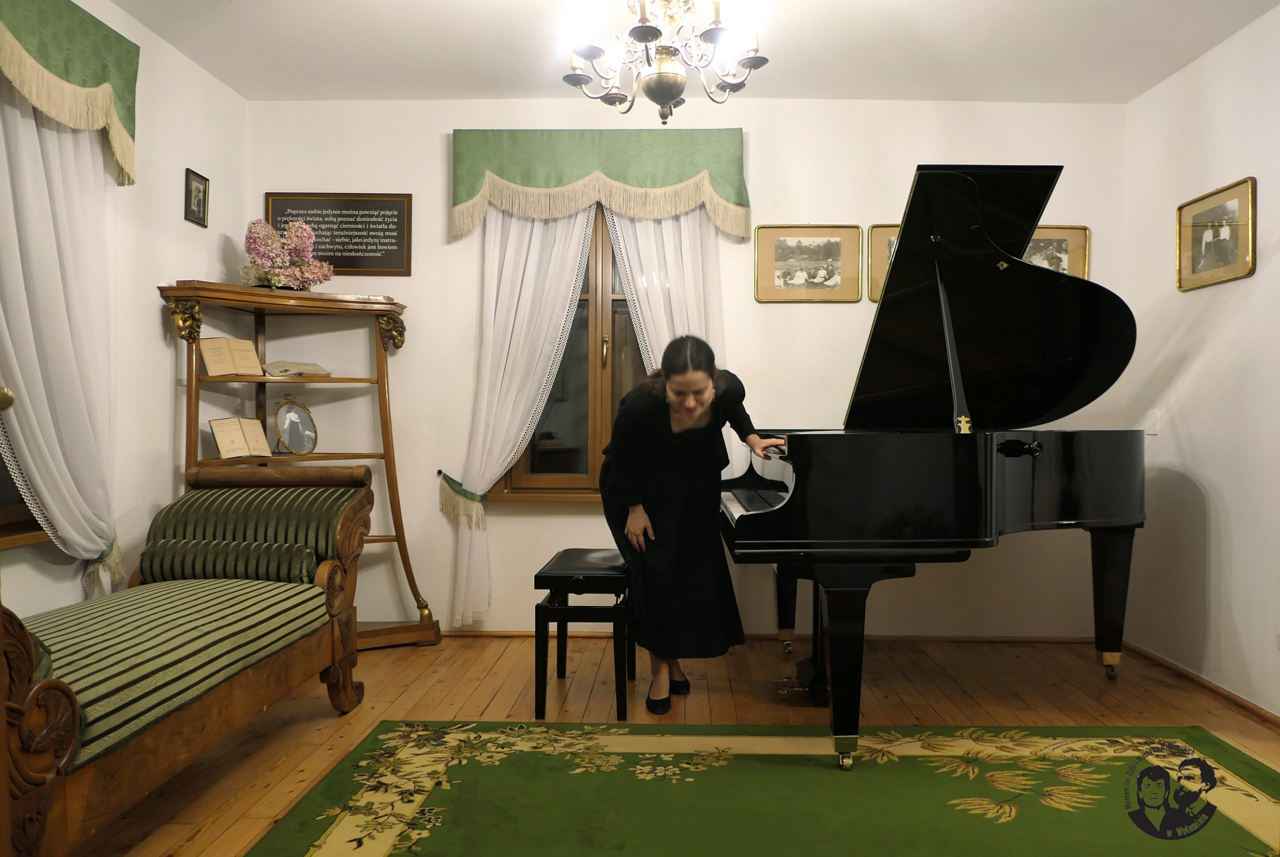 Fotografia kolorowa przedstawiająca artystkę w ukłonie, lewa ręka na instrumencie, widać cały fortepian oraz wnętrze salonu „Domu nad Łąkami”.