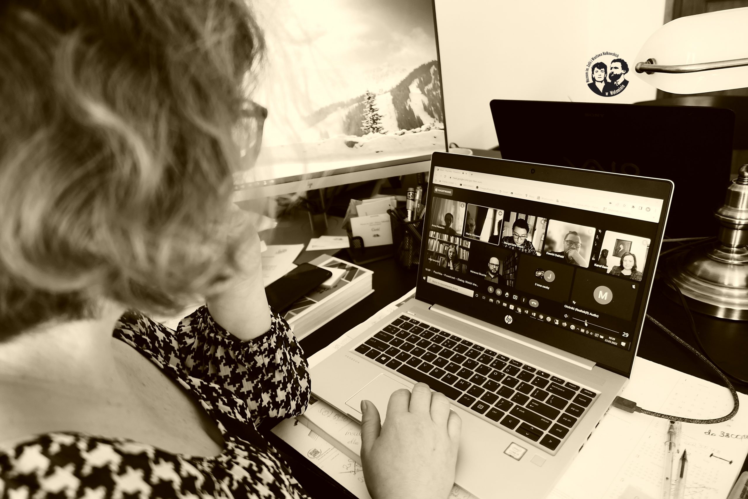 Fotografia w odcieniach sepii, sytuacyjna, na zdjęciu widoczny monitor komputera z osobami biorącymi udział w panelu oraz pracownik muzeum