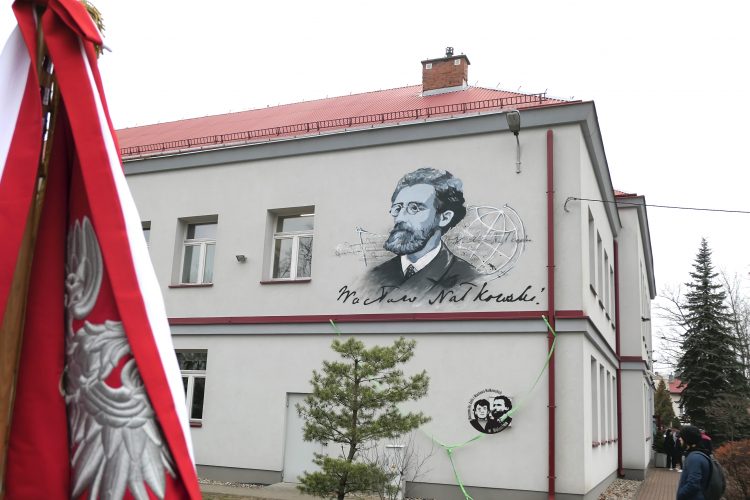 Fotografia kolorowa, pozioma, przedstawiająca widok ściany budynku szkoły, na piętrze której znajduje się mural z podobizną Wacława Nałkowskiego oraz jego podpisem i schematycznym przedstawieniem globusa. Z lewej strony fotografii fragment sztandaru szkoły.